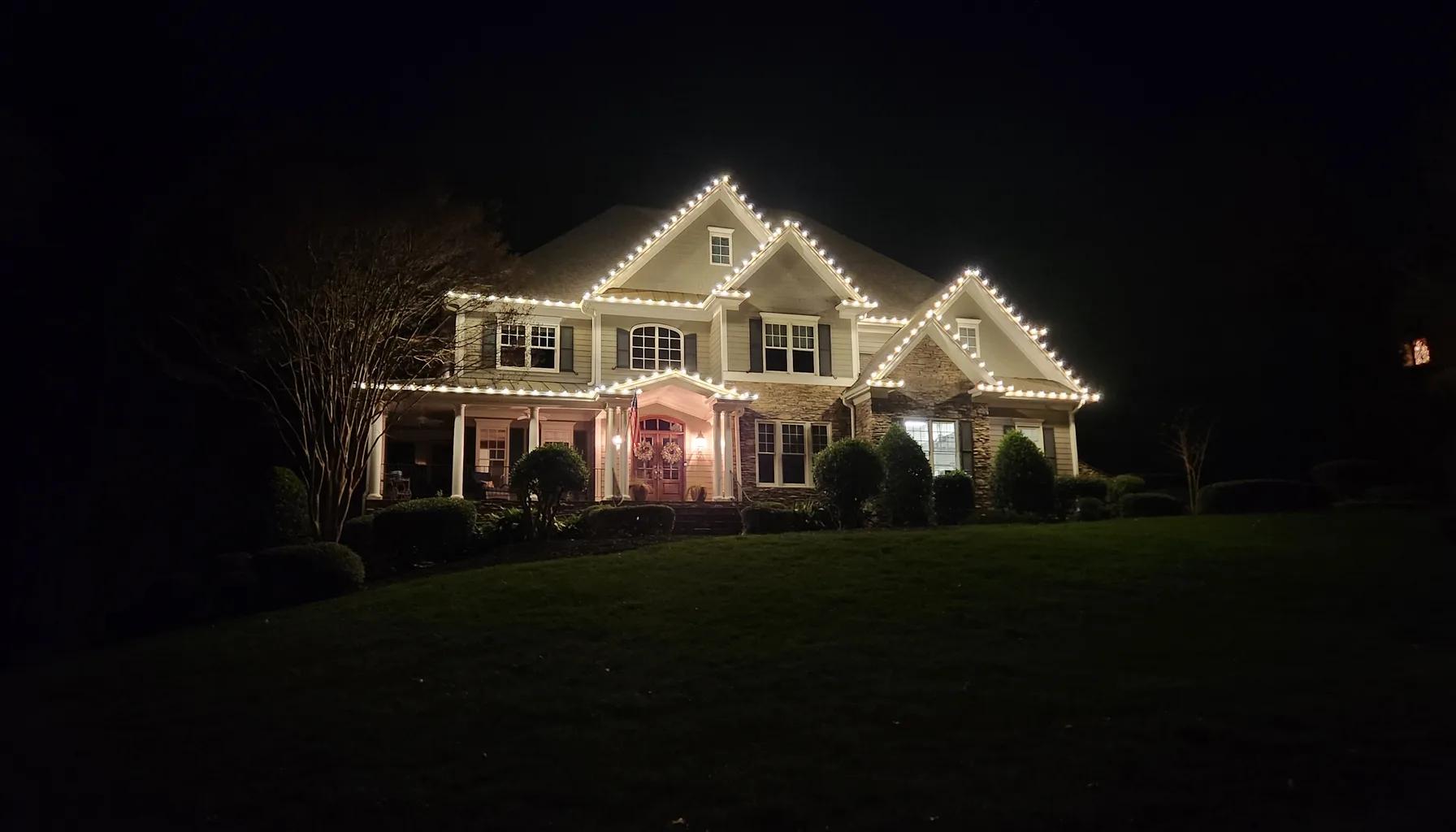 Two-story home near Sunset Lake in Holly Springs, NC with warm white C9 Christmas lights outlining every roofline and gable, and lit wreaths on the front door